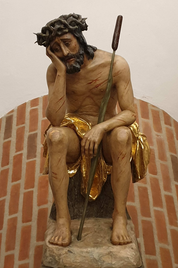 Skulptur von Jesus, der auf einem Block sitzt und traurig blickend seinen Kopf stützt 