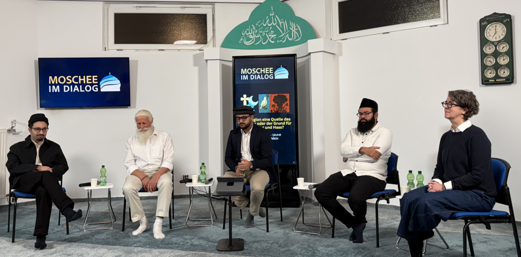 Gesamtansicht der Podiumsteilnehmer. Im Hintergrund sind auf Bildschirmen der Schriftzug "Moschee im Dialog und der Titel der Veranstaltung zu lesen.