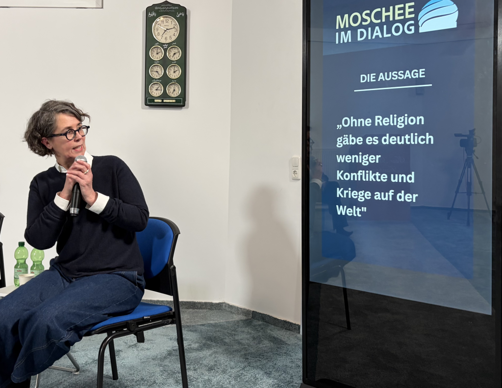 Johanna Steinbach, rechts daneben ein Bildschirm mit der ersten These "Ohne Religion gäbe es deutlich weniger Konflikte und Kriege auf der Welt"